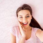 Self top above view portrait of pretty, brunette, charming, cute, glad, pleased, positive girl holding palm near cheek looking at camera, shooting selfie on front camera, lying on back on bed